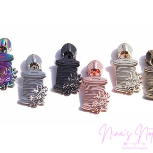 May include: Assortment of six zipper pulls in various colors, each featuring a detailed spool of thread with a floral design. The colors include iridescent, black, silver, rose gold, gold, and antique brass.