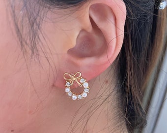 May include: Gold tone earrings with a pearl and rhinestone wreath design. The wreath has a small bow in the center.