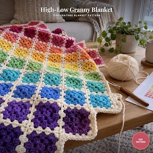 May include: A crochet blanket featuring a rainbow-colored granny square pattern with white borders. The text High-Low Granny Blanket Temperature Blanket Pattern is visible. A ball of white yarn and a wooden crochet hook are nearby.