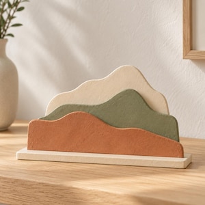 Könnte beinhalten: Ein dekoratives Tischobjekt bestehend aus drei geschichteten, bergförmigen Ausschnitten in Creme, Salbeigrün und Terrakotta-Orange. Die Formen sind auf einer rechteckigen cremefarbenen Basis montiert und erzeugen einen Tiefeneffekt.