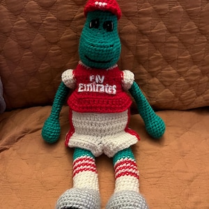 May include: A handmade green and red crocheted dinosaur wearing a red hat and a red and white soccer jersey with the text "Fly Emirates". The dinosaur has gray shorts and striped socks.