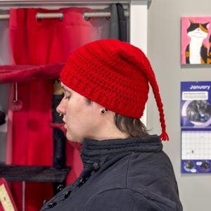 May include: A bright red, handmade crochet hat with a long, braided tassel. The hat is a pointed style, likely a gnome or elf hat. The hat is made of a thick yarn, and the texture of the crochet is visible.