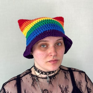 Könnte beinhalten: Ein gehäkelter Hut in Regenbogenfarben mit Katzenohren. Der Hut zeigt ein lebendiges Farbspektrum, darunter Rot, Orange, Gelb, Grün, Blau und Lila. Der Hut ist im Bucket-Stil mit breiter Krempe.