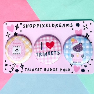 May include: A pink card with three circular badges. The badges display a Tamagotchi-style character, the text "I ❤️ TRINKETS", and a bear in a hat. The card is decorated with hearts and stars. The text "TRINKET BADGE PACK" is also visible.