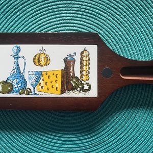 May include: A wooden serving board with a white tile inset featuring a still life of a cheese wedge, a bottle of oil, a pepper grinder, a bunch of olives, and a green pepper. The tile is surrounded by a dark brown wooden frame.