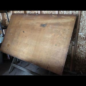 May include: A large wooden drafting table with a metal frame. The table is made of plywood and has a worn surface. The table is sitting on a wooden floor.