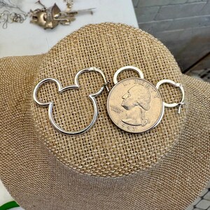May include: Silver-toned Mickey Mouse-shaped hoop earrings displayed next to a U.S. quarter coin. The earrings are on a tan burlap surface, with other jewelry and a teacup visible in the background.