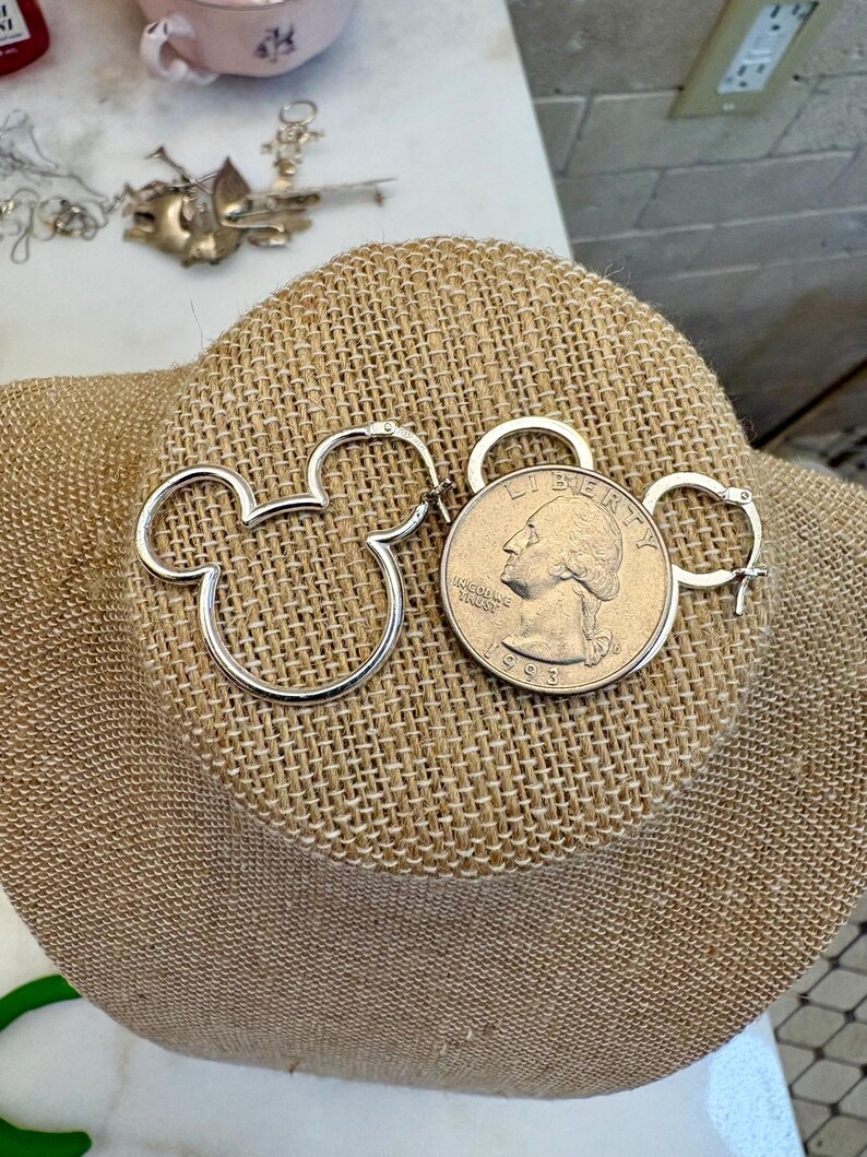 May include: Silver-toned Mickey Mouse-shaped hoop earrings displayed next to a U.S. quarter coin. The earrings are on a tan burlap surface, with other jewelry and a teacup visible in the background.