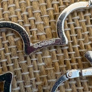May include: Close-up of silver-toned hoop earrings with a Mickey Mouse head shape. The word "DISNEY" is stamped on the earring. The earrings are on a textured, tan surface.