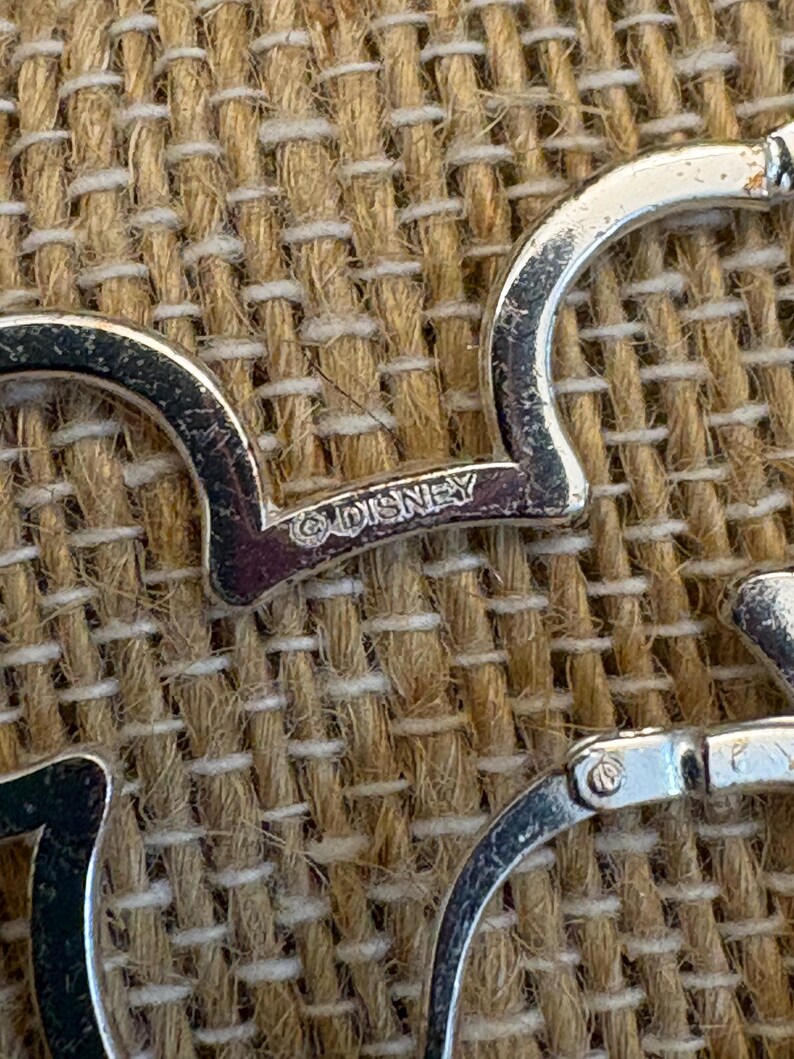 May include: Close-up of silver-toned hoop earrings with a Mickey Mouse head shape. The word "DISNEY" is stamped on the earring. The earrings are on a textured, tan surface.