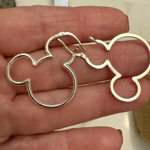 May include: A pair of silver-colored hoop earrings in the shape of Mickey Mouse's head. The earrings are made of a shiny metal and have a simple, minimalist design. The earrings are shown against a neutral background.