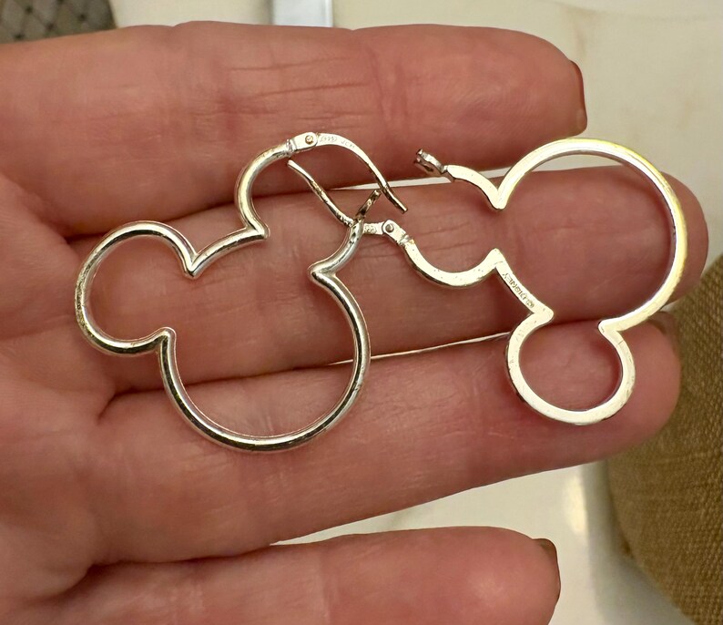 May include: A pair of silver-colored hoop earrings in the shape of Mickey Mouse's head. The earrings are made of a shiny metal and have a simple, minimalist design. The earrings are shown against a neutral background.