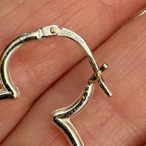 May include: Close-up of a pair of silver hoop earrings. The earrings are heart-shaped and have a hinged closure. The metal is stamped with "925" and "JCM".