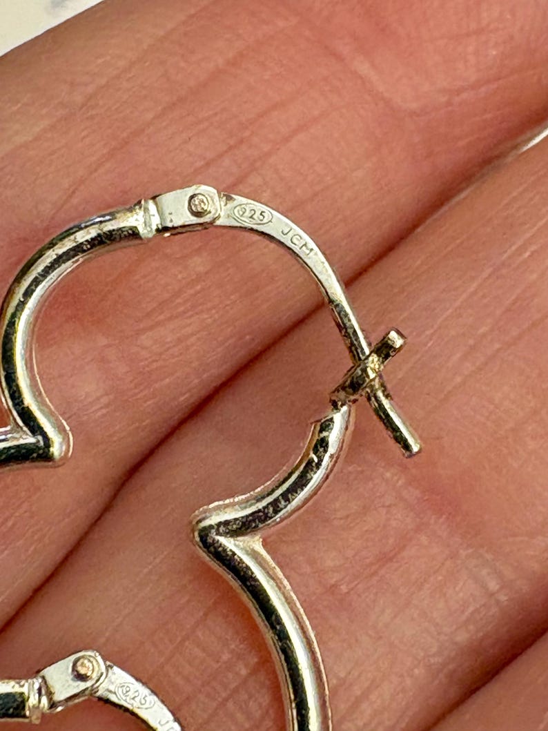 May include: Close-up of a pair of silver hoop earrings. The earrings are heart-shaped and have a hinged closure. The metal is stamped with "925" and "JCM".