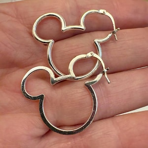 May include: A pair of silver-colored hoop earrings in the shape of Mickey Mouse's head. The earrings are made of a shiny metal and have a classic design. The earrings are a fun and playful accessory.