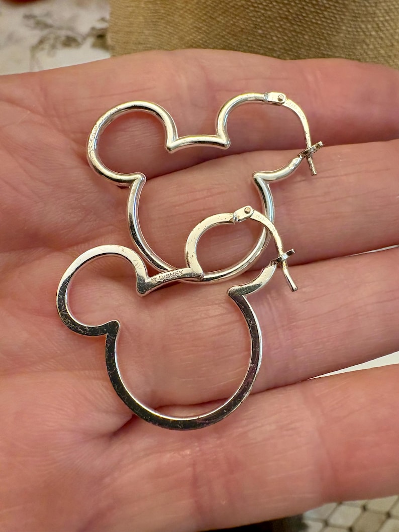 May include: A pair of silver-colored hoop earrings in the shape of Mickey Mouse's head. The earrings are made of a shiny metal and have a classic design. The earrings are a fun and playful accessory.