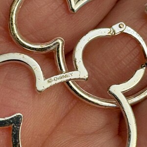 May include: Close-up of silver-colored Mickey Mouse-shaped earrings. The earrings are made of a shiny metal and feature the Disney logo. The design is a classic and recognizable silhouette.