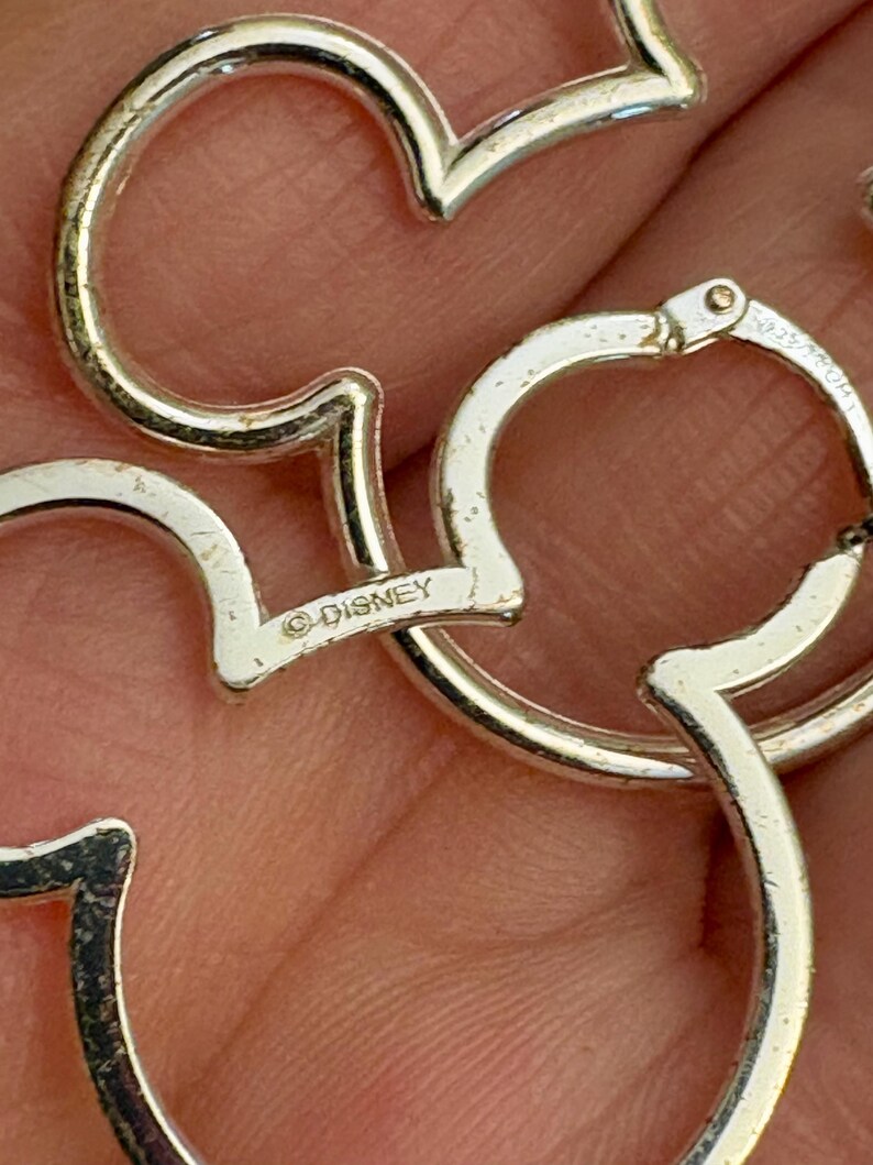 May include: Close-up of silver-colored Mickey Mouse-shaped earrings. The earrings are made of a shiny metal and feature the Disney logo. The design is a classic and recognizable silhouette.