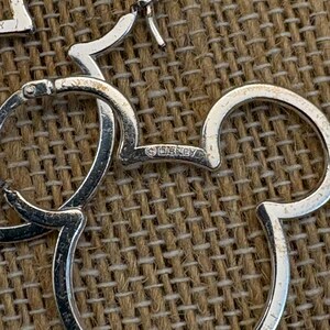 May include: Silver-toned Mickey Mouse hoop earrings. The earrings are shaped like Mickey Mouse's head and ears. The word "&copy;DISNEY" is visible on one of the earrings. The earrings are on a textured, brown surface.