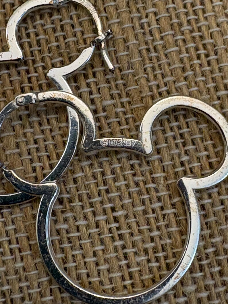 May include: Silver-toned Mickey Mouse hoop earrings. The earrings are shaped like Mickey Mouse's head and ears. The word "&copy;DISNEY" is visible on one of the earrings. The earrings are on a textured, brown surface.