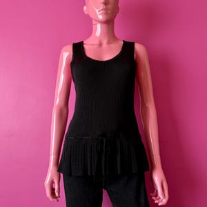 May include: Black ribbed tank top with a pleated peplum hem and drawstring detail. The top is displayed on a mannequin against a pink background. The tank top is sleeveless and has a scoop neckline.