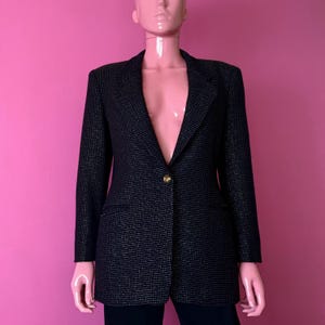 May include: A black blazer with a subtle dotted pattern and a single gold button closure. The blazer has a classic notched lapel and two front pockets. The garment is displayed on a mannequin against a pink background.