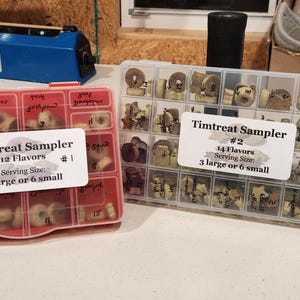 May include: Two clear plastic containers filled with small, round, dog treats. The containers are labeled "Timtreat Sampler" with the number of flavors and serving size. The container on the left is red and the container on the right is clear.