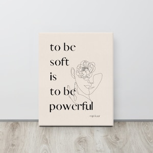 Peut inclure: Une impression sur toile beige avec un dessin au trait noir d'un visage de femme avec une fleur couvrant un œil. Le texte "to be soft is to be powerful" est imprimé en noir sous le dessin. La citation est attribuée à "rupi kaur" en caractères plus petits.