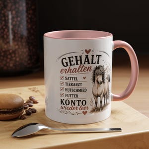 以下が含まれることがあります： 内側と持ち手が淡いピンク色の白いセラミック製マグカップ。馬のイラストと、Gehalt erhalten、Sattel、Tierarzt、Hufschmied、Futter、Konto wieder leerというドイツ語のテキストがプリントされており、小さな赤いハートで装飾されています。