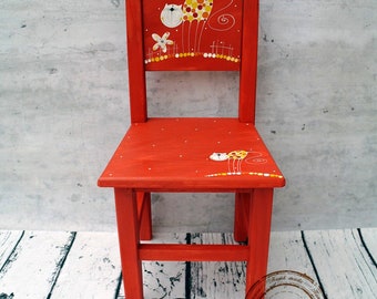 Puede incluir: Una silla de madera roja para niños con un diseño pintado a mano de un gato con manchas amarillas y naranjas. La silla tiene un fondo blanco con acentos amarillos y naranjas.