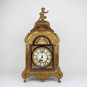 Könnte beinhalten: Eine vergoldete Kaminuhr mit einer Cherubfigur auf der Oberseite. Das Zifferblatt der Uhr hat römische Ziffern und einen weißen Hintergrund mit floralen Akzenten. Die Uhr ist mit komplizierten floralen Mustern in Gold, Rot und Schwarz verziert.