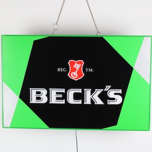 Peut inclure: Une enseigne verte et noire avec le texte "BECK'S" en lettres blanches. L'enseigne a un bouclier rouge avec une clé à l'intérieur et le texte "REG. TM."