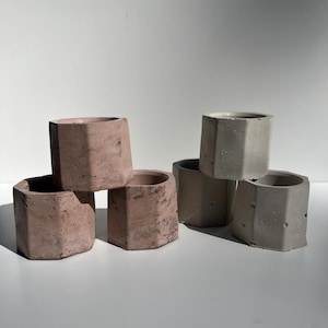 May include: A collection of six small, hexagonal concrete planters in shades of pink and gray. The planters have a rough, textured surface and are arranged in a group, showcasing their geometric design. Ideal for small plants or succulents.