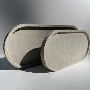 Może przedstawiać: Dwie owalne tace betonowe w różnych rozmiarach. Tace są w kolorze jasnoszarym z teksturowaną powierzchnią. Większa taca znajduje się za mniejszą, tworząc efekt warstwowy. Odpowiednie do dekoracji wnętrz.