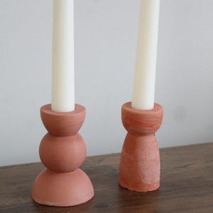 May include: Two terracotta-colored candle holders with white candles. One holder has a stacked sphere design, the other a tapered shape. The candle holders are on a wooden surface.