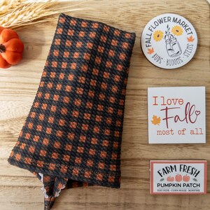 May include: A black and orange checkered fabric item with a white lace trim, alongside decorative wooden signs. The signs read "Fall Flower Market," "I love Fall most of all," and "Farm Fresh Pumpkin Patch."