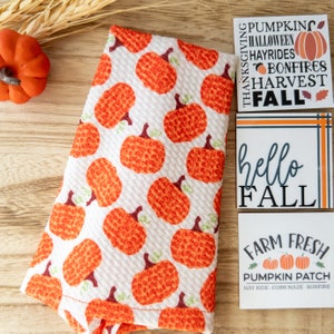 May include: White fabric with a repeating pattern of orange pumpkins with green stems. The image also includes three small signs with fall-themed text, including "Hello Fall" and "Farm Fresh Pumpkin Patch."