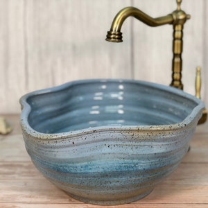 Könnte beinhalten: Ein blaues Keramikwaschbecken mit einem wellenförmigen Rand und einer gesprenkelten Glasur. Das Waschbecken ist so konzipiert, dass es wie eine Muschel aussieht. Ein goldener Wasserhahn ist im Hintergrund sichtbar.