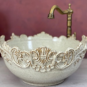 May include: A white ceramic bathroom sink with an ornate, decorative rim. The sink is round and has a speckled finish.