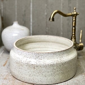 May include: A white ceramic vessel sink with a speckled finish. The sink is round and has a wide, flat rim. A gold faucet is mounted to the wall behind the sink.