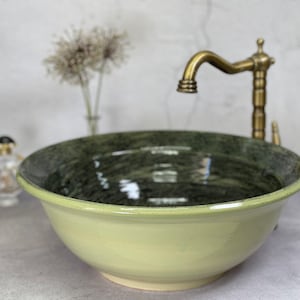 Könnte beinhalten: Ein runder, hellgelber Keramikwaschtisch mit dunkelgrünem Innenraum. Das Waschbecken steht vor einem neutralen Hintergrund mit einem Messingwasserhahn. Dekorative Elemente sind eine kleine Parfümflasche und eine Vase mit Trockenblumen.