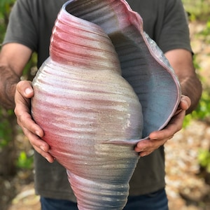 May include: A large ceramic sculpture in the shape of a seashell. The shell is painted in shades of pink, blue, and white, with a speckled texture.