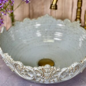 Puede incluir: Un lavabo de cer&aacute;mica azul claro con un borde decorativo y ornamentado. El lavabo tiene un acabado moteado y un desag&uuml;e dorado.