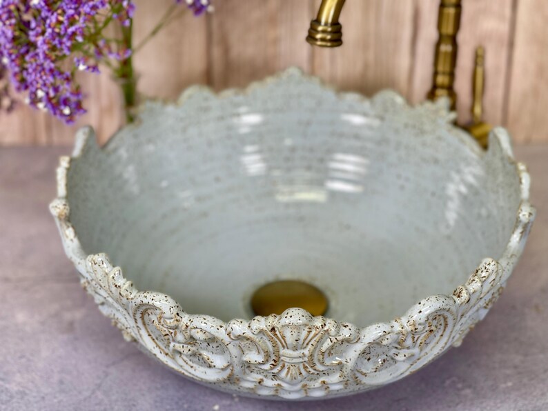 Puede incluir: Un lavabo de cer&aacute;mica azul claro con un borde decorativo y ornamentado. El lavabo tiene un acabado moteado y un desag&uuml;e dorado.