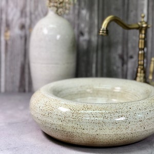May include: A speckled, off-white ceramic sink with a rounded shape. A vintage-style brass faucet is visible on the right. A matching vase with dried flowers is in the background, creating a neutral, spa-like aesthetic.