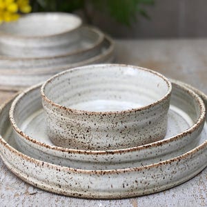 May include: A collection of handmade ceramic bowls and plates. The off-white tableware has a speckled texture and a brown rim. The items are arranged on a light-coloured surface, showcasing the rustic design.