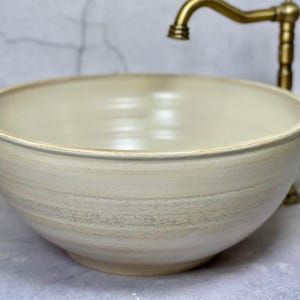 May include: A round, cream-colored ceramic vessel sink with a smooth, matte finish and subtle horizontal banding. The basin features a wide, open top and a tapered base, accompanied by a vintage-style, antique brass faucet with a curved spout.