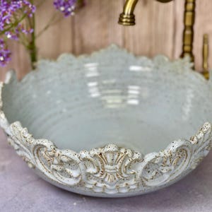 Puede incluir: Un lavabo de cer&aacute;mica azul claro con un borde decorativo y ornamentado. El lavabo es redondo y tiene un acabado liso y brillante.