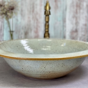May include: A white ceramic bowl with a speckled glaze and a wide, slightly flared rim. The bowl is sitting on a light gray surface.