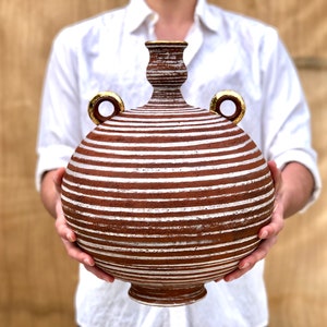 May include: A large, round, brown ceramic vase with white stripes and gold accents. The vase has two handles and a narrow neck.